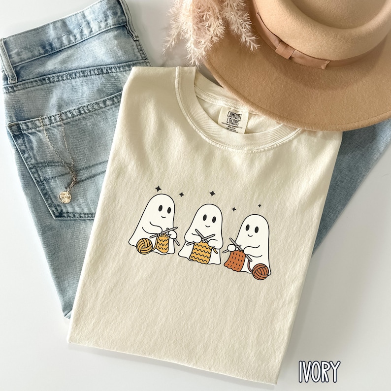 May include: Ivory-colored t-shirt featuring a graphic of three cartoon ghosts knitting with yarn. The shirt is laid on top of blue jeans, a tan hat, and a silver necklace. The text "IVORY" is printed at the bottom of the shirt.