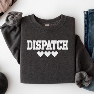 May include: A dark charcoal gray crewneck sweatshirt featuring the word DISPATCH printed in bold white capital letters, positioned above three white heart icons. The garment has long sleeves with ribbed cuffs and a classic round neckline.