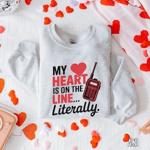 May include: A gray sweatshirt with a red heart and a walkie-talkie graphic. The text on the sweatshirt reads "My heart is on the line... Literally."