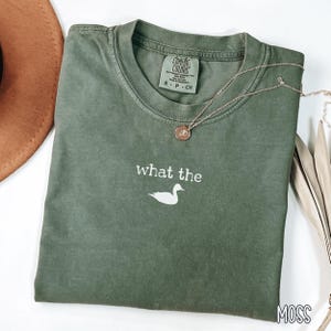May include: Moss green t-shirt with the phrase "what the" and a duck graphic. The shirt has a crew neck and short sleeves. A gold necklace with a pendant is draped across the shirt.