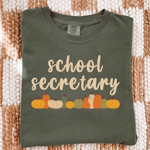 May include: An olive green t-shirt featuring the text "school secretary" in a light tan script font. Below the text is a row of various pumpkins and gourds in shades of orange, cream, and muted green. The shirt has a Comfort Colors brand label.