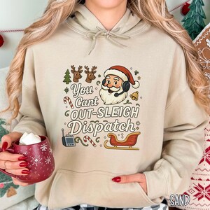 May include: A sand-colored hoodie featuring a festive graphic with Santa Claus wearing a headset, a sleigh, and the text "You Can't Out-Sleigh Dispatch." The design includes reindeer, candy canes, and a bell. A red mug with white snowflake designs is held in the hand.