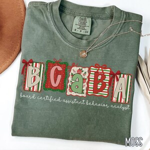 May include: A moss green t-shirt with the letters "BCABA" in a gift box design. The text "board certified assistant behavior analyst" is below the letters. A gold necklace with a pendant is on the shirt.