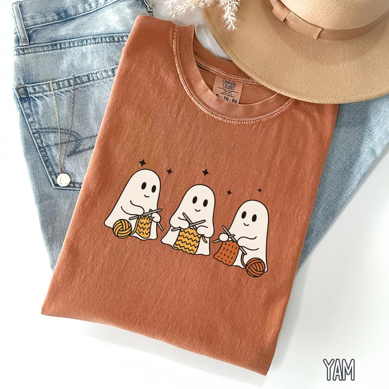 Yam-colored shirt with three cute cartoon ghosts knitting with yarn in yellow and orange tones, surrounded by stars.