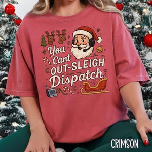 May include: Crimson-colored t-shirt with a festive graphic. The design features Santa Claus wearing a headset, reindeer, a sleigh, and the text "You Can't Out-Sleigh Dispatch." Candy canes and a radio are also included in the design.