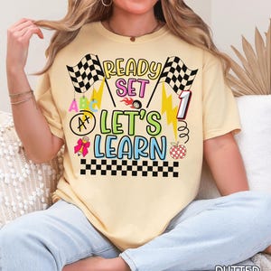 May include: Butter-colored t-shirt with the text "READY SET LET'S LEARN" in colorful, playful lettering. The design includes checkered flags, lightning bolts, and an apple. The shirt is a casual, short-sleeved style.