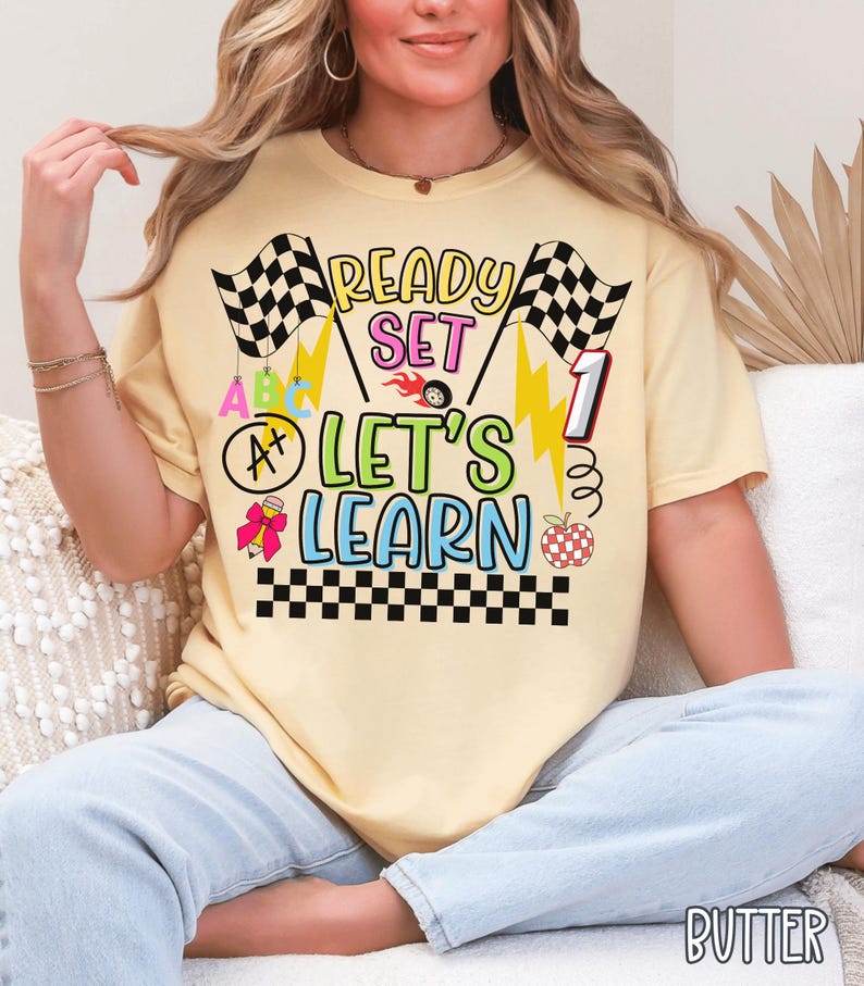 May include: Butter-colored t-shirt with the text "READY SET LET'S LEARN" in colorful, playful lettering. The design includes checkered flags, lightning bolts, and an apple. The shirt is a casual, short-sleeved style.