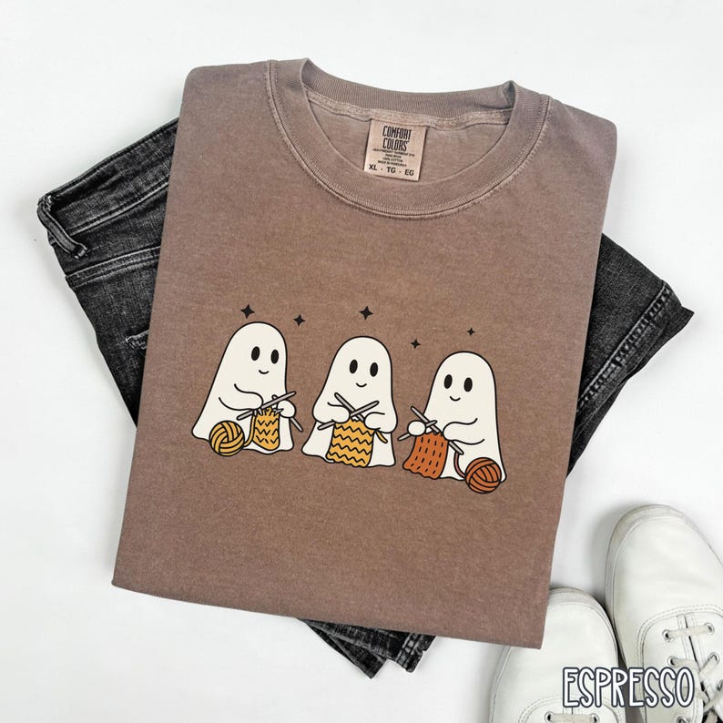 May include: Espresso-colored Comfort Colors t-shirt featuring a graphic of three cartoon ghosts knitting with yarn. The shirt is paired with black denim jeans and white sneakers. The tag reads XL - TG - EG.