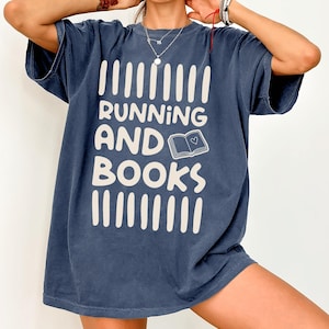 May include: A navy blue short-sleeve t-shirt featuring the cream-colored text RUNNING AND BOOKS, with a small graphic of an open book containing a heart symbol, framed by vertical cream-colored bars above and below the text.