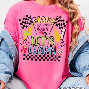 May include: A bright pink t-shirt with the words "READY SET LET'S LEARN" in a colorful, playful font. The design includes checkered flags, lightning bolts, an apple, and a grade of A+. The shirt is a comfortable fit.