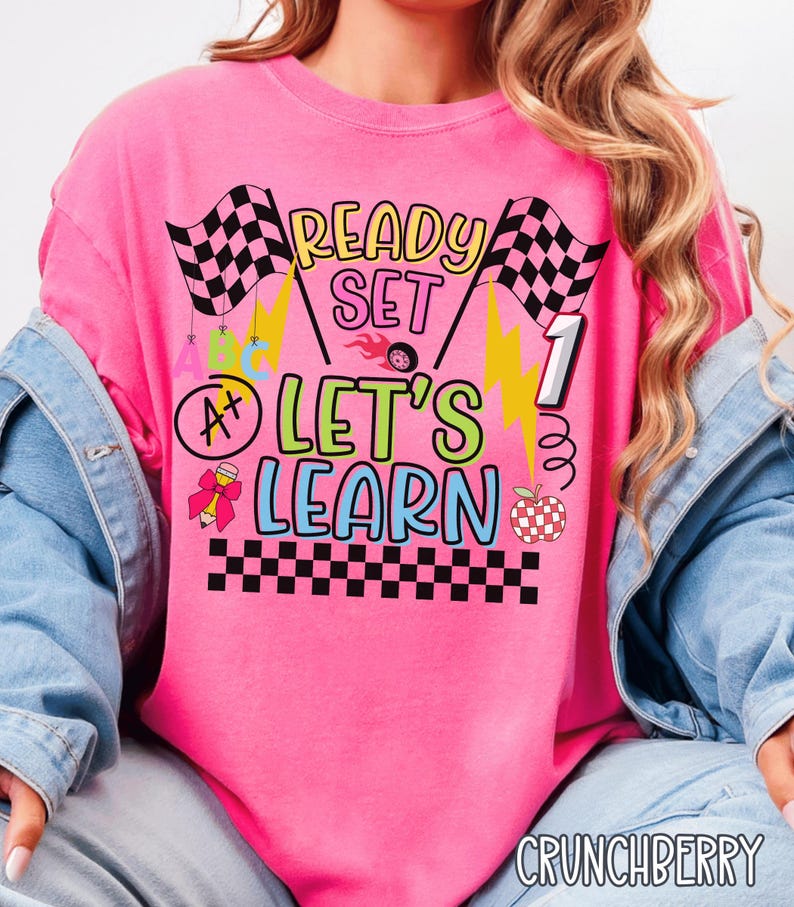 May include: A bright pink t-shirt with the words "READY SET LET'S LEARN" in a colorful, playful font. The design includes checkered flags, lightning bolts, an apple, and a grade of A+. The shirt is a comfortable fit.