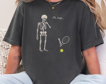 Può includere: Una t-shirt grigio antracite con la grafica di uno scheletro con un braccio staccato che tiene una racchetta da tennis. Accanto allo scheletro c'è una pallina da tennis gialla. Il testo "Oh, snap..." è stampato in bianco sopra la spalla.