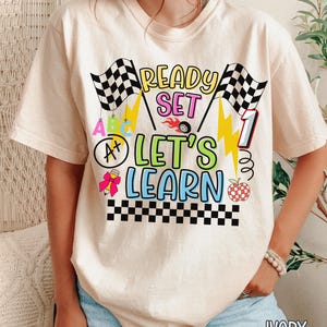 Cream colored teacher t-shirt featuring colorful “Ready Set Let’s Learn” text with racing flags, lightning bolts, apples, and classroom themed graphics for first grade teachers.