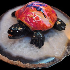 Puede incluir: Una pequeña figurita de tortuga pintada a mano con un diseño abstracto colorido en su caparazón. La tortuga está sentada sobre una rebanada de ágata blanca con un borde negro y marrón.