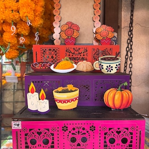 May include: A three-tiered wooden display stand featuring vibrant colors and cut-out designs. The levels are decorated with illustrations of sugar skulls, marigolds, candles, a taco, a pumpkin, and a mug. The bottom tier is bright pink.