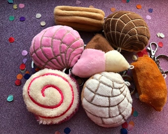 Può includere: Una collezione di portachiavi in peluche a forma di pane dolce messicano, tra cui conchas rosa, marroni e bianche, un rotolo e un pane rettangolare arancione. Su una superficie glitterata viola con coriandoli.