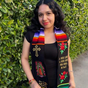 May include: A graduation stole with a colorful striped design and black panels. The stole features embroidered roses, a cross, and an image of the Virgin Mary. Text on the stole reads "Class of 2026" and a Spanish quote.