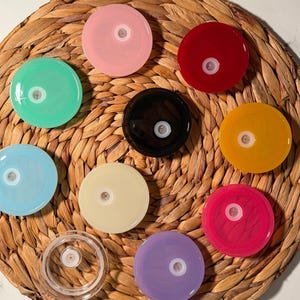 May include: A collection of colorful, round lids in various shades, including pink, green, red, yellow, and purple. Each lid has a central white component. The lids are arranged on a woven, circular mat.