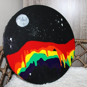 Puede incluir: Una pintura circular con fondo negro y un arcoíris de colores que gotean desde la parte superior. El arcoíris está formado por rojo, naranja, amarillo, verde, azul y morado. Hay una luna blanca y estrellas en el fondo.