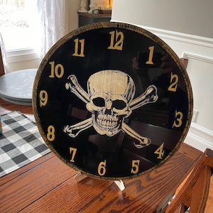 Könnte beinhalten: Eine schwarze und goldene Uhr mit einem Totenkopf- und Knochenkreuz-Design. Das Zifferblatt der Uhr hat römische Ziffern und ist vor einem schwarzen Hintergrund platziert. Der Totenkopf und die Knochenkreuze sind golden.