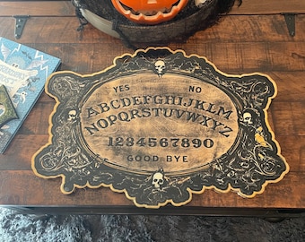 Könnte beinhalten: Ein schwarzes und goldenes Ouija-Brett mit den Buchstaben des Alphabets, den Zahlen 1-9 und 0 sowie den Wörtern "YES", "NO" und "GOOD BYE".