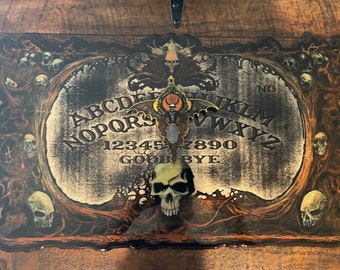 May include: A black and white Ouija board with a skull in the center. The board is surrounded by a dark, ornate frame with skulls and vines. The letters A-Z, numbers 1-9, and the words "GOODBYE" and "NO" are printed on the board.