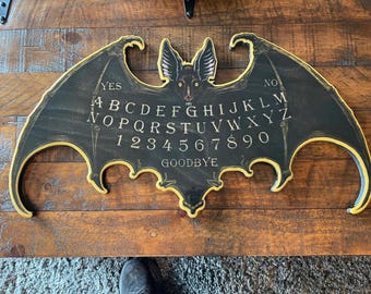 May include: A black wooden bat-shaped Ouija board with a yellow border and a bat illustration in the center. The board features the letters A-Z, numbers 1-9, 0, and the word "Goodbye". The words "Yes" and "No" are printed on the board.