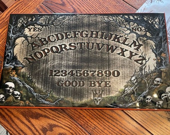 May include: A black and white Ouija board with a spooky forest scene. The board features the alphabet, numbers 1-9 and 0, and the words "YES", "NO", and "GOOD BYE".