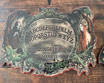 May include: A wooden Ouija board with a black and white alphabet, numbers 1-9 and 0, and the word "Goodbye". The board is surrounded by a dark green and black border with images of a skeletal figure and a woman with long red hair.