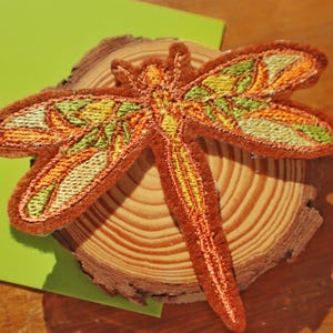 May include: An embroidered dragonfly patch featuring a stained-glass style pattern with vibrant orange, yellow, and green thread colours. The insect design has a detailed segmented body and four wings with intricate stitching on a brown felt base.