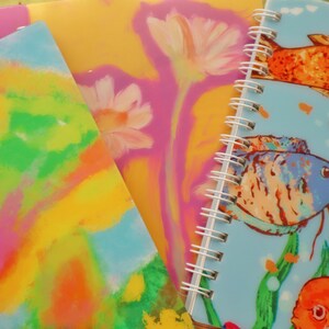 Puede incluir: Una colección de cuadernos en espiral coloridos. Uno presenta una cubierta azul claro con ilustraciones de peces. Otro tiene un diseño floral amarillo y rosa. El tercero tiene un diseño abstracto amarillo, verde y azul.
