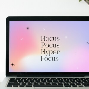 Op de afbeelding: Een laptop met een roze en paarse gradiënt achtergrond op het scherm. De tekst "Hocus Pocus Hyper Focus" wordt in zwarte tekst op het scherm weergegeven.