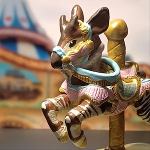 May include: A brown and white zebra carousel figure with a light blue bridle and saddle blanket. The zebra is attached to a gold-colored vertical pole. The figure features painted details in pink, gold, and light blue, with a textured mane and hooves.