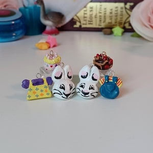 May include: A collection of miniature, hand-painted charms. The charms include two white bunny faces, a yellow dress with green flowers, blue overalls, a cupcake, and a chocolate dessert. The charms are designed for jewelry making or crafting.
