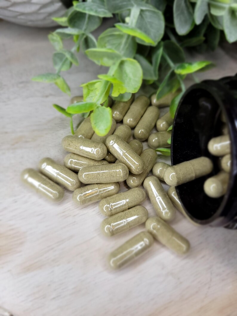May include: A scattering of green capsules spilling from a black container, with some capsules on a light surface. Green leafy sprigs are in the background. The capsules appear to be filled with a green powder, suggesting a herbal supplement or medication.