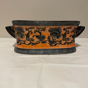 以下が含まれることがあります： 2つの取っ手が付いた楕円形のセラミックプランター。プランターはオレンジ色の背景に黒と緑の花柄で飾られています。プランターは黒と灰色の縁取りがされています。