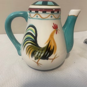 Könnte beinhalten: Eine weiße Teekanne aus Keramik mit einem türkisfarbenen Henkel und Ausguss. Die Teekanne ist mit einem bunten Hahn-Design und einem blauen, roten und schwarzen Muster auf dem Deckel verziert.