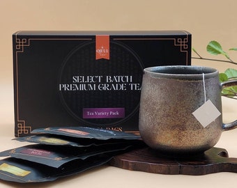 May include: A black box with the text "Select Batch Premium Grade Tea" and "Tea Variety Pack" on it. The box is next to a brown ceramic mug with a tea bag hanging from the handle. The mug is on a wooden coaster.