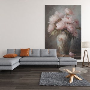 Op de afbeelding: Een grijze hoekbank met twee oranje kussens staat voor een schilderij van roze pioenen in een vaas. Een glazen salontafel met een houten onderstel staat op een grijs tapijt voor de bank.