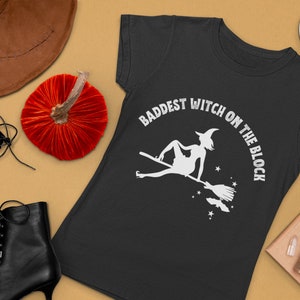 May include: Black t-shirt with a white graphic of a witch flying on a broomstick. The text "Baddest Witch on the Block" is written in a white circle around the witch.