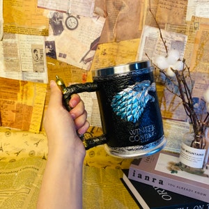 May include: A black and silver mug with a faux leather exterior and a silver handle. The mug features a blue and silver wolf head with the words "Winter is Coming" printed below it.