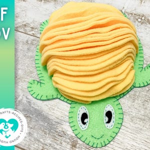 Könnte beinhalten: Eine grüne Filzturtel mit einer gelben Filzschale. Die Schildkröte hat ein lächelndes Gesicht mit zwei schwarzen Augen. Die Schale besteht aus mehreren Schichten Filz, was ihr ein strukturiertes Aussehen verleiht.