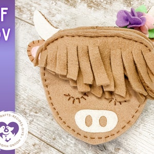 Könnte beinhalten: Ein brauner Filz-Highland-Cow-Kopf mit weißer Nase und Augen. Die Kuh hat einen braunen Filzfransen als Haar und eine lila Blume auf dem Kopf.