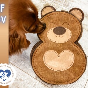 Könnte beinhalten: Ein braunes Filzspielzeug für Hunde in Form eines Bären mit einem Herzen auf der Brust. Der Bär hat eine braune Nase und Augen. Ein Hund beschnuppert das Spielzeug.