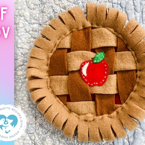 Könnte beinhalten: Ein braunes Filz-Apfelkuchen-Hundespielzeug mit einem roten Filzapfel und einem grünen Filzblatt. Die Tortenkrusten sind aus braunen Filzstreifen gefertigt und haben einen ausgefransten Rand.