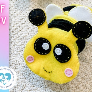 May include: A yellow felt bee with black stripes, white wings, and black eyes. The bee has a stitched smile and pink cheeks. The bee is lying on a beige surface.