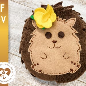 Könnte beinhalten: Ein brauner Filz-Igel mit einer gelben Filzblume auf dem Kopf. Der Igel hat gestickte Augen, Nase und Mund. Der Igel ist ein Plüschtier.