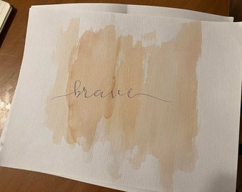 May include: A white card with a light brown watercolor wash and the word "brave" written in black ink.