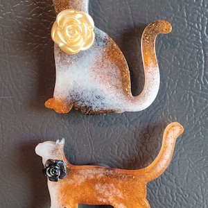 Puede incluir: Dos figuritas de gato de resina con detalles de purpurina. Un gato es blanco con manchas naranjas y negras y tiene una rosa dorada en la espalda. El otro gato es blanco con manchas naranjas y tiene una rosa negra en la espalda.