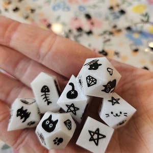 May include: A set of seven white dice with black cat-themed illustrations. The dice are in a hand, and each die has a different design, including a cat, a skull, a bone, a star, a diamond, a heart, and a paw print.